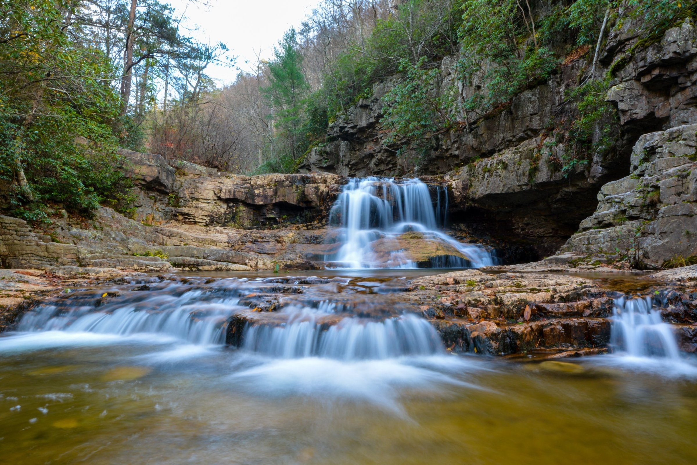 Long-exposure waterfall spilling over layered rock into a still amber pool, framed by forest