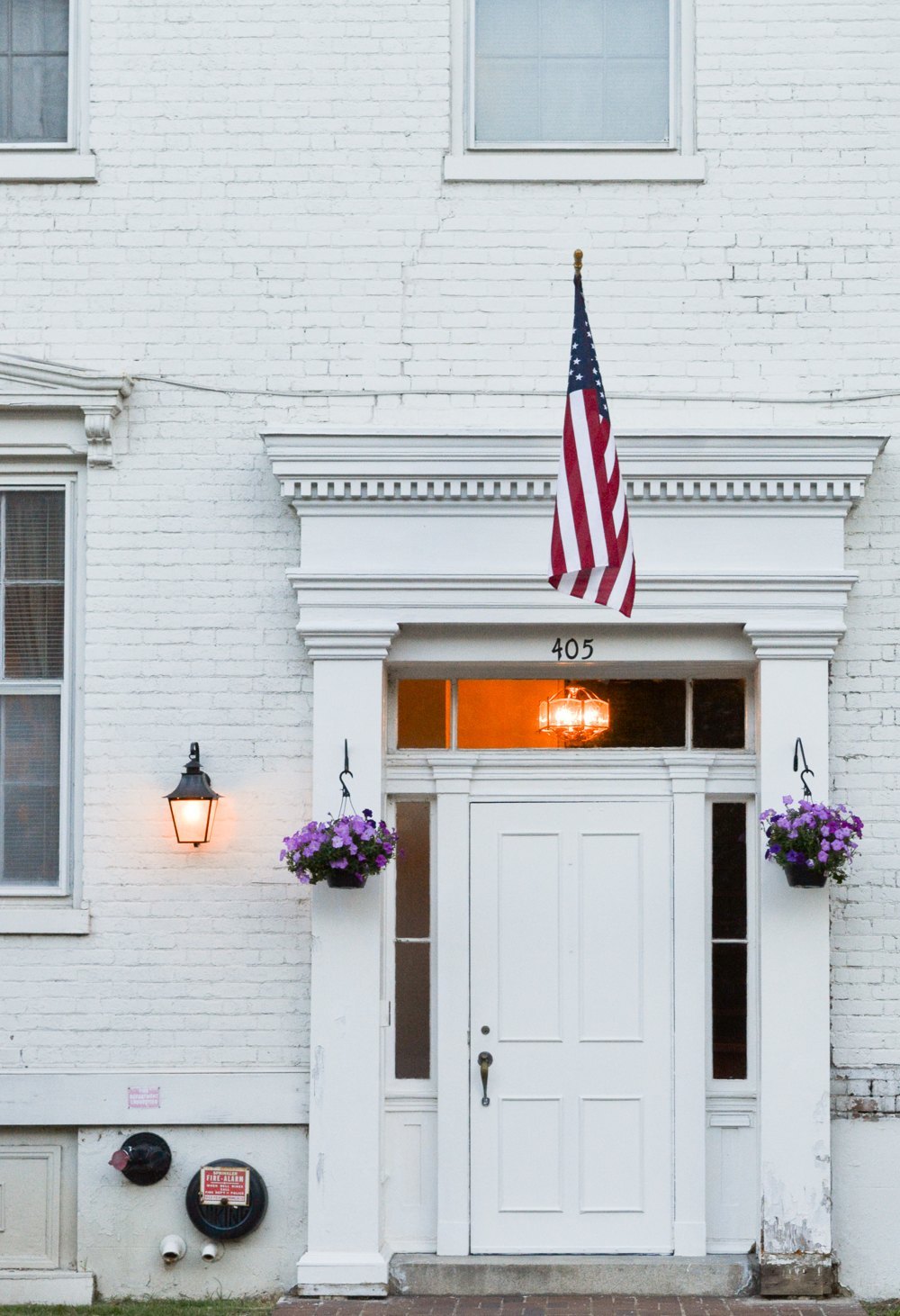 White brick house front at dusk, an American flag over the lit doorway and baskets of purple flowers either side