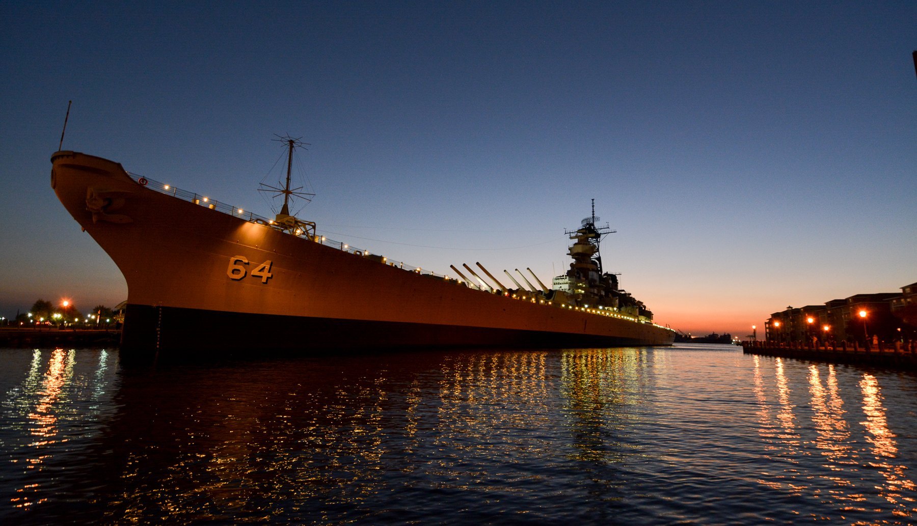Battleship Wisconsin lit up at its pier after sunset, lights reflected in the water