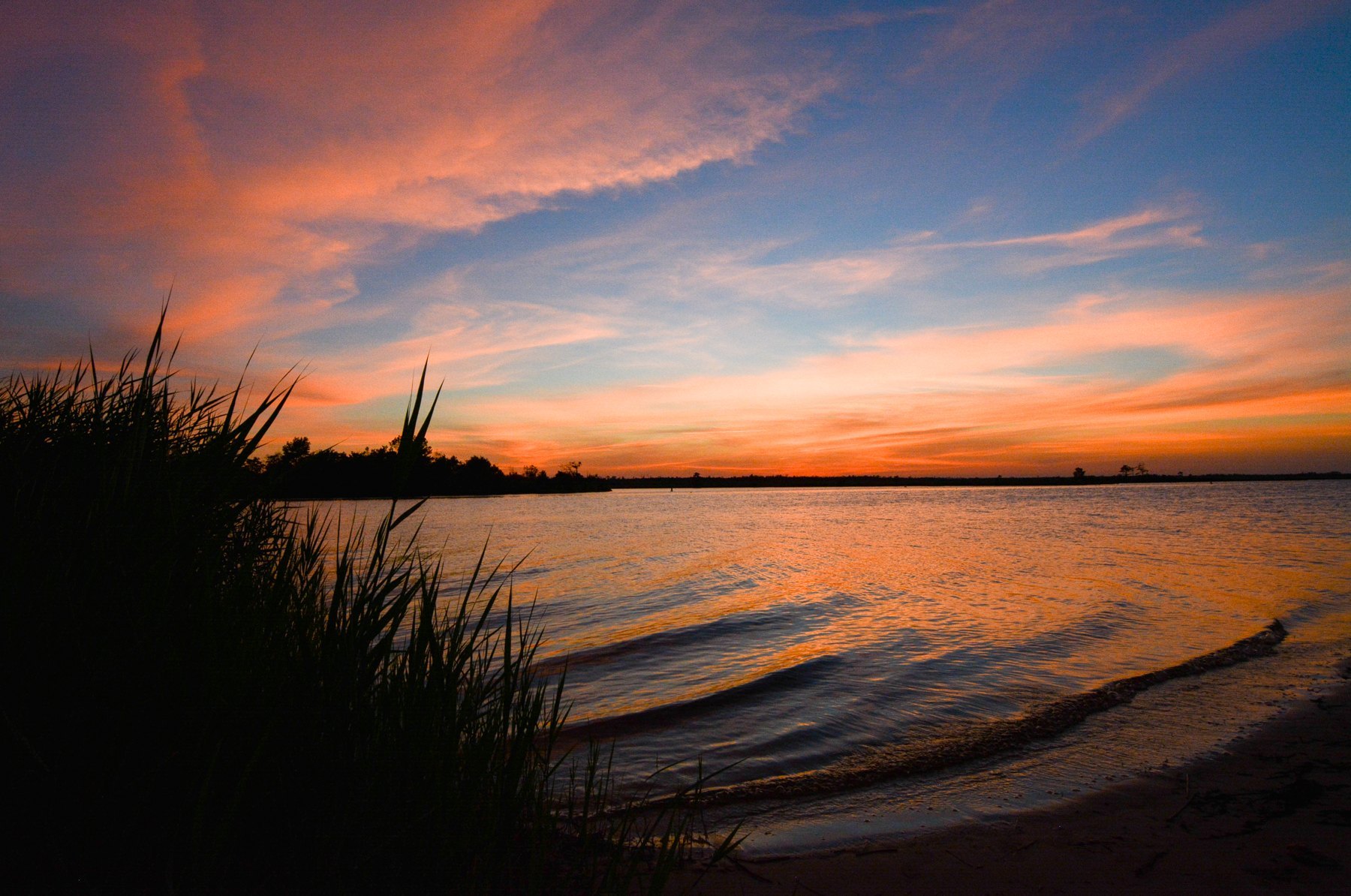 Sunset over a wide river, pink and orange clouds above and marsh grass silhouetted in the foreground