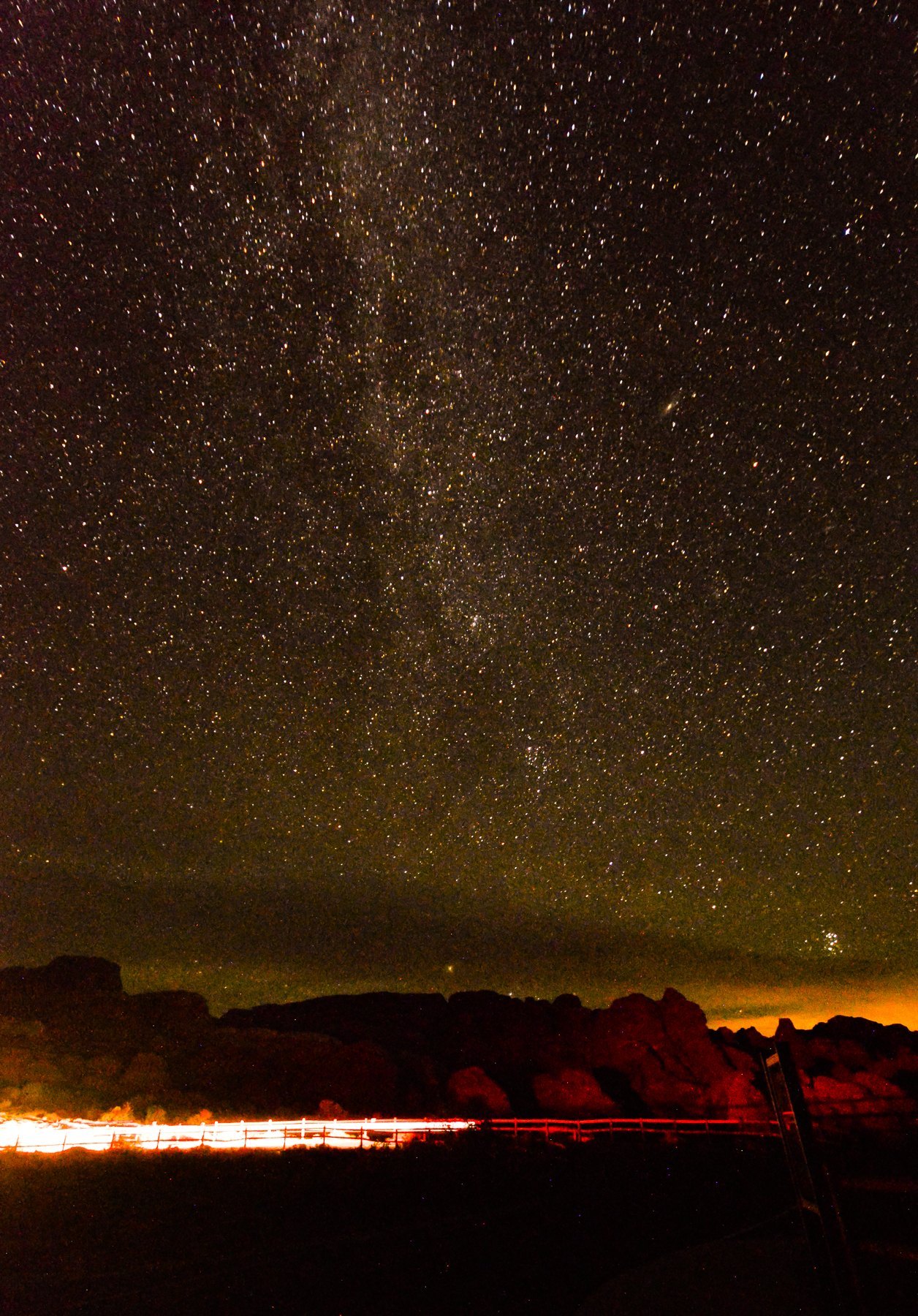 Star-filled sky over red rock, a passing car's light trail glowing along a fence line